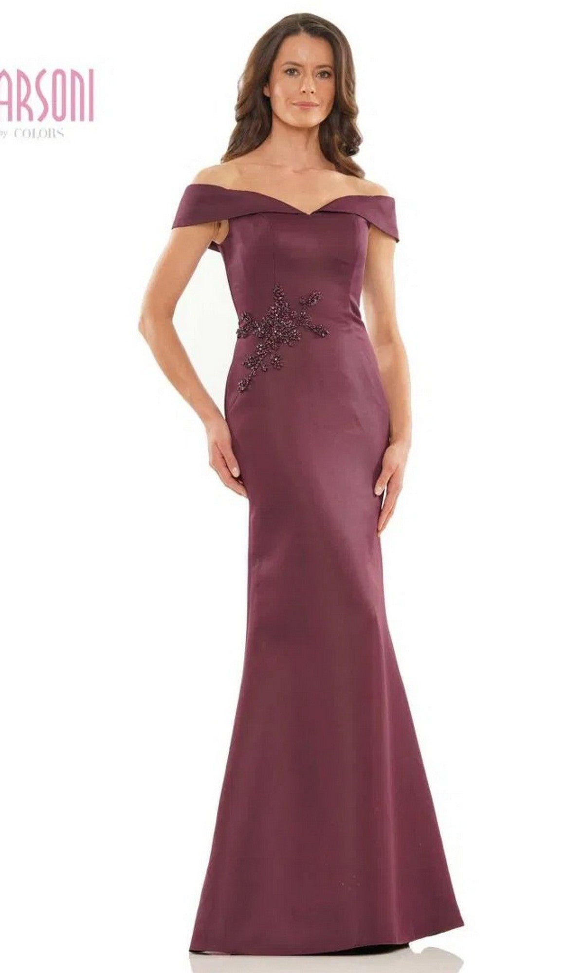 A woman in the Marsoni MV1003 long formal dress in wine, featuring an off-the-shoulder satin design and beaded floral embellishment at the left waist, stands against a plain background.